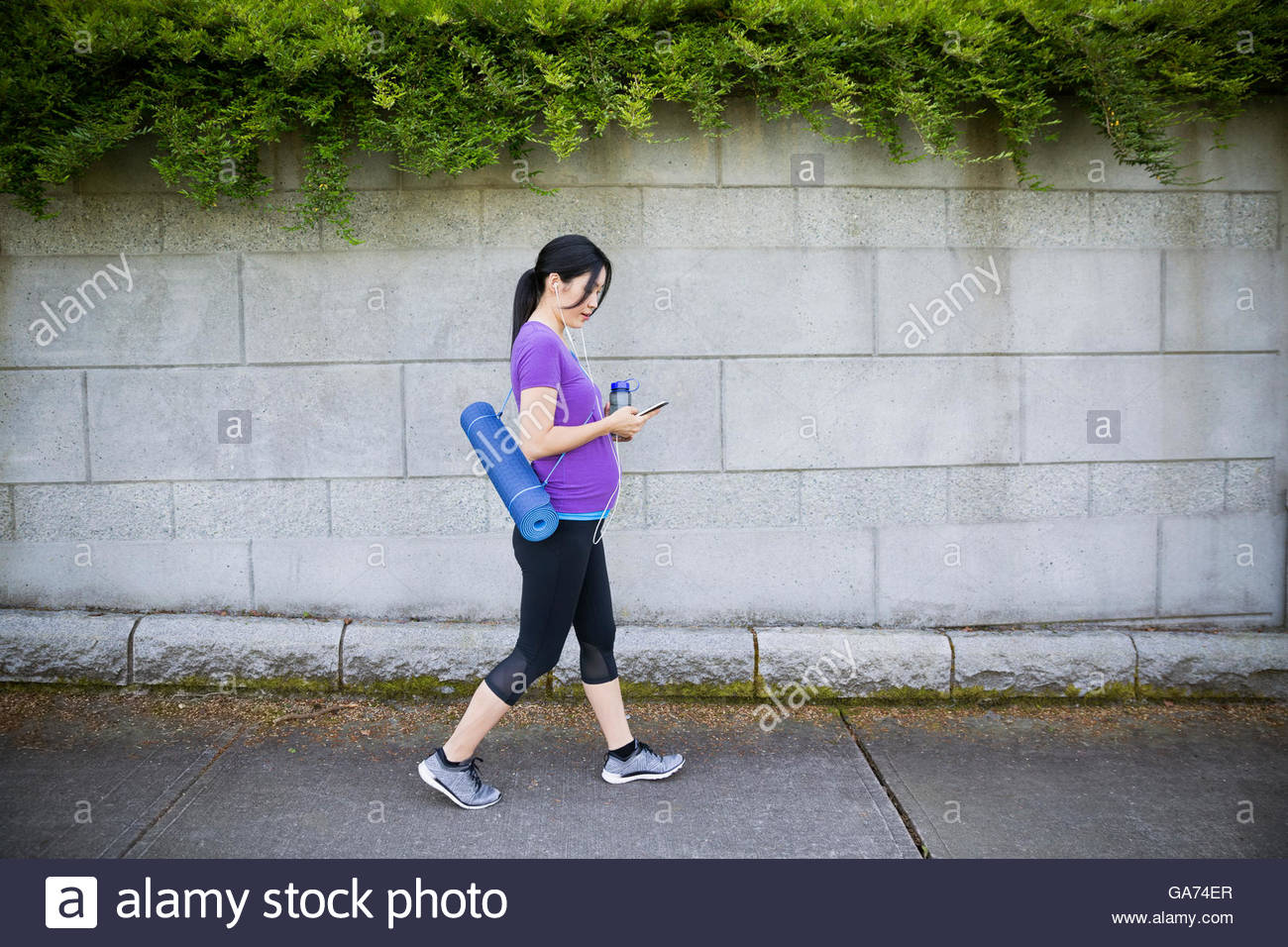 Pregnant woman with yoga mat and headphones using cell phone on sidewalk Stock Photo