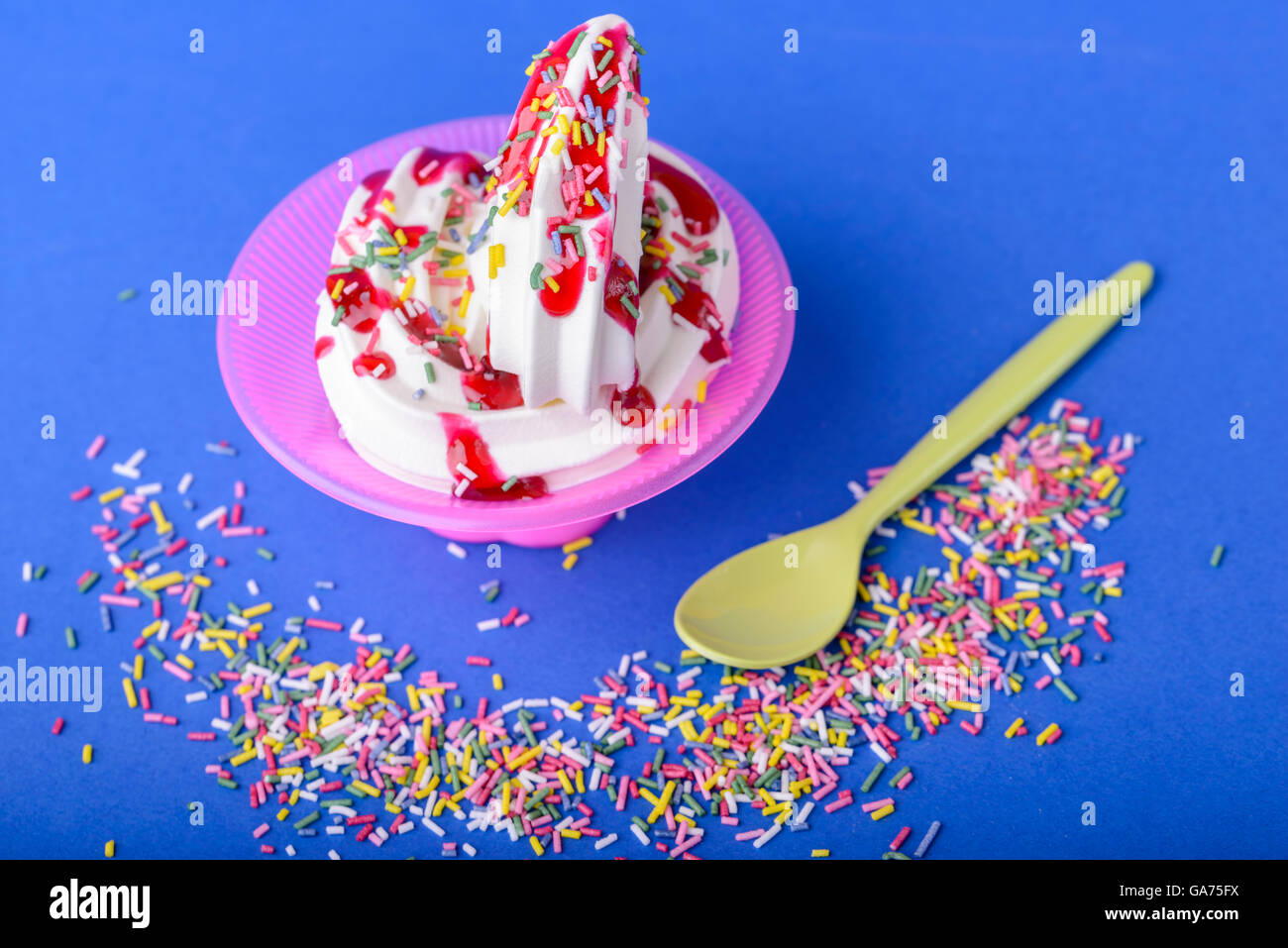 frozen yogurt with black cherry topping and rainbow sprinkles on blue background with yellow spoon and rainbow sprinkles Stock Photo
