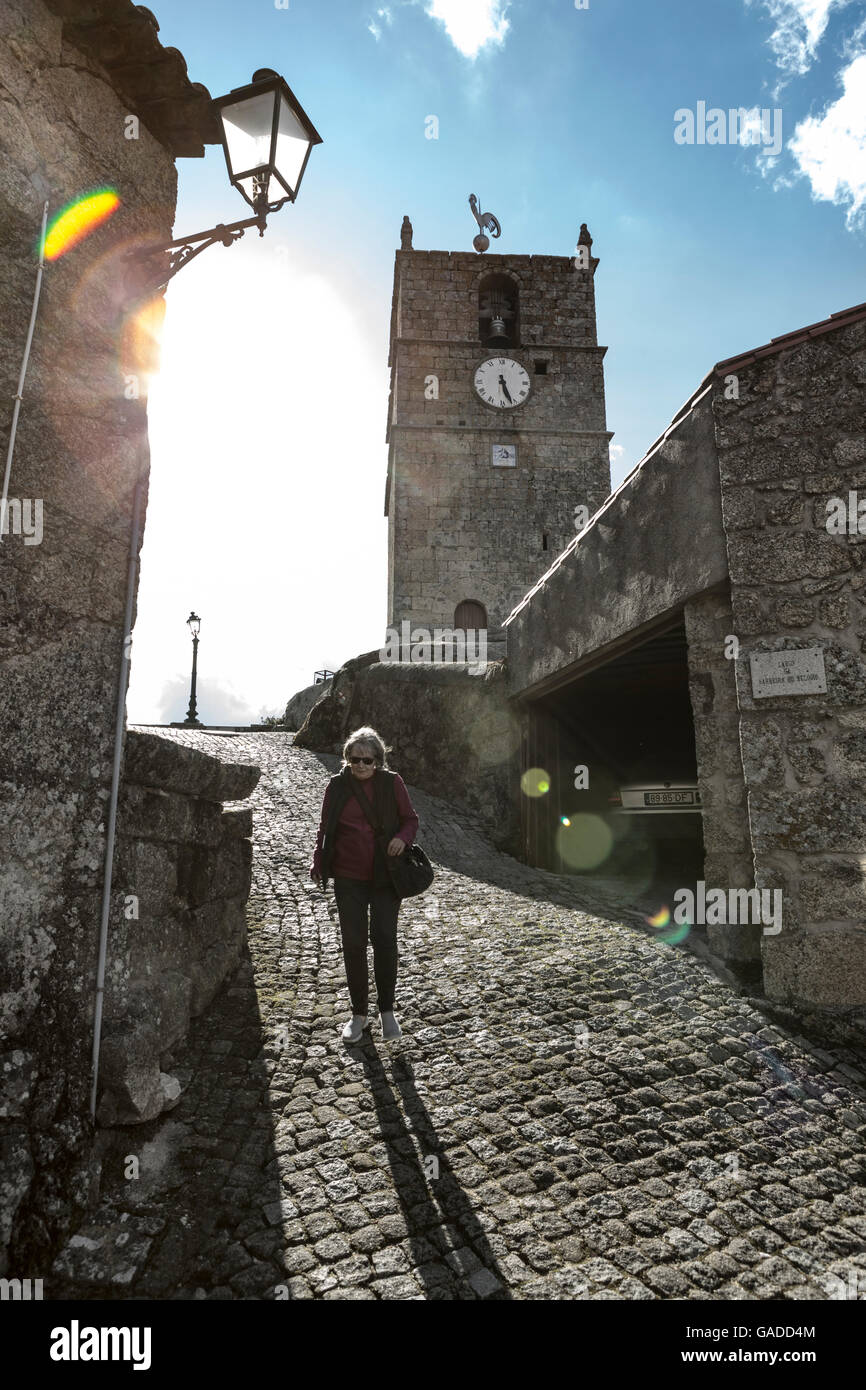 A woman walking down a cobbled street in an ancient village in Europe with a church clocktower behind, against the light with sun flare, high contrast Stock Photo
