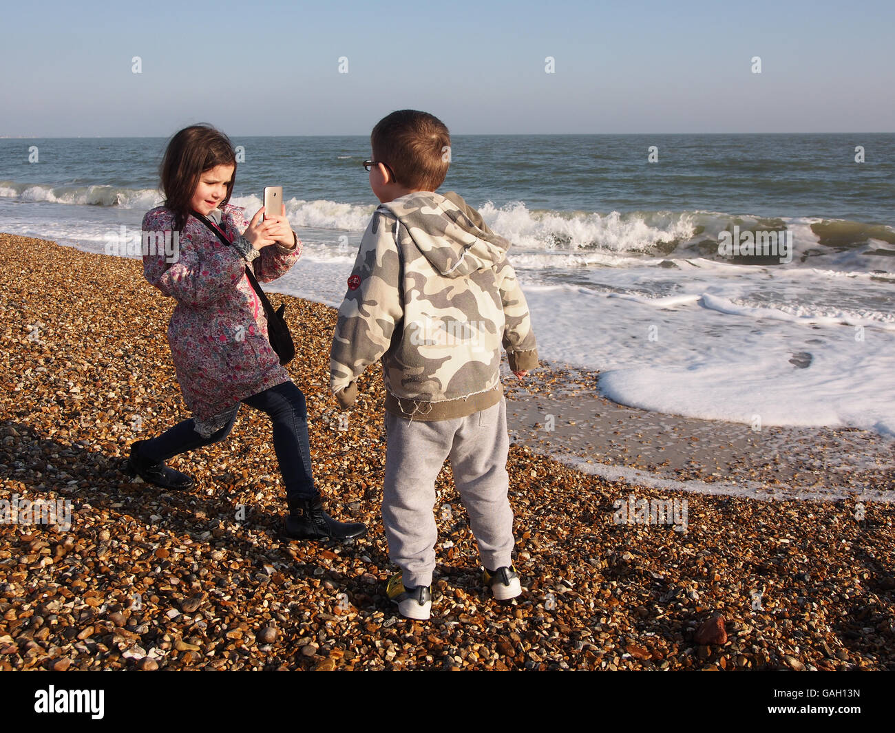 A young girl taking photographs of her brother on her smart phone on the beach Stock Photo