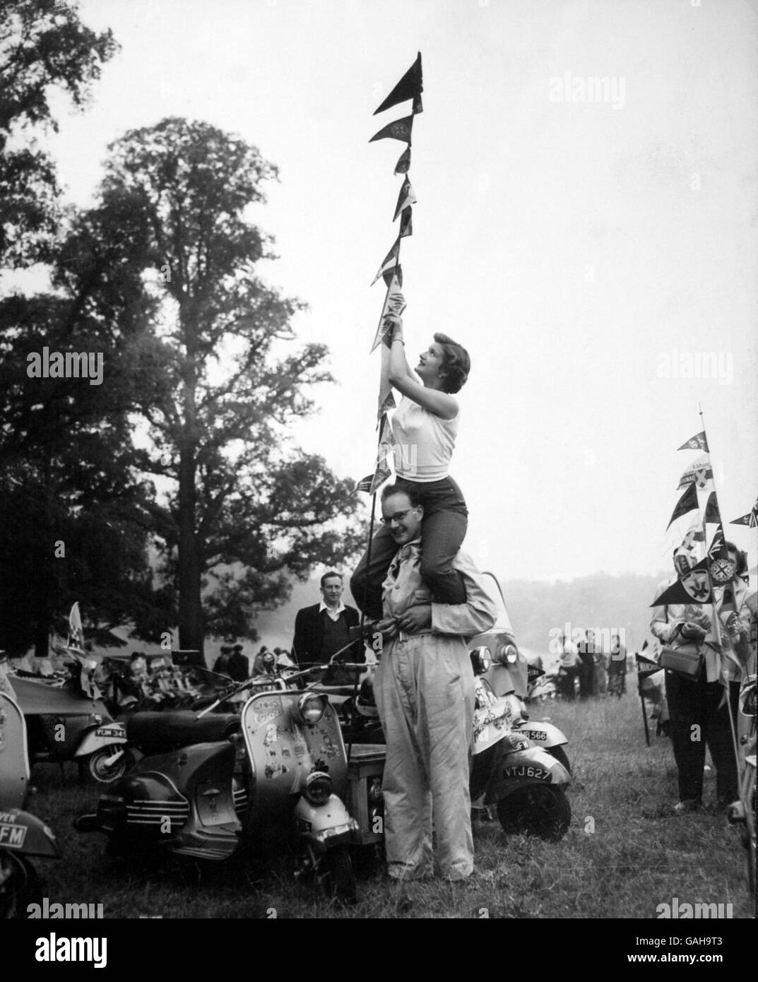 Muriel Patterson perched on Carl Watsons shoulders fixes one of the pennants on his scooter. The pennants represent the 14 countries that Carl has been to on his Vespa. Stock Photo