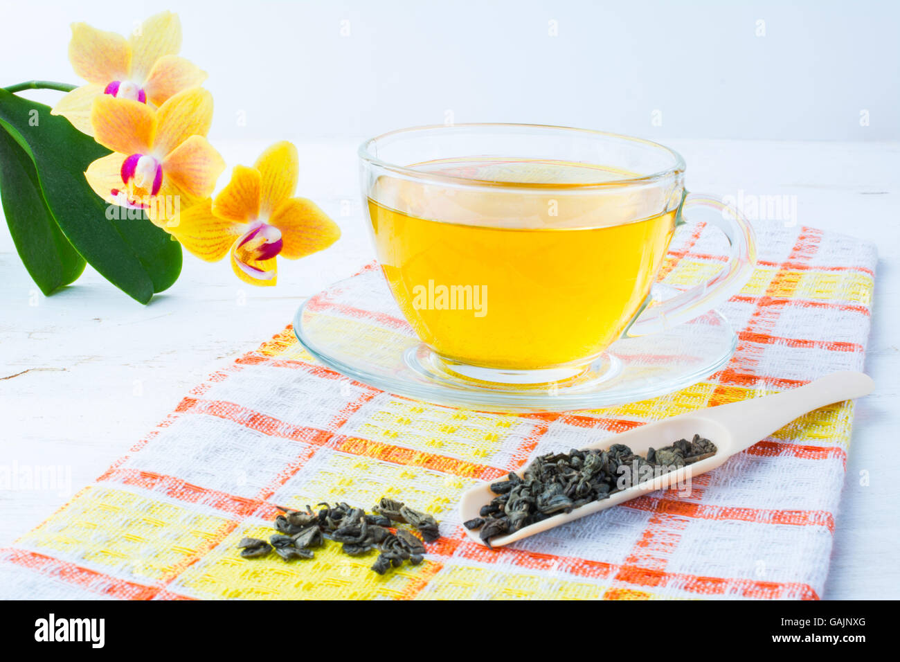 Cup of green tea on orange checkered napkin with a yellow orchid on a white wooden background. Cup tea. Cap of tea. Tea.  Green Stock Photo
