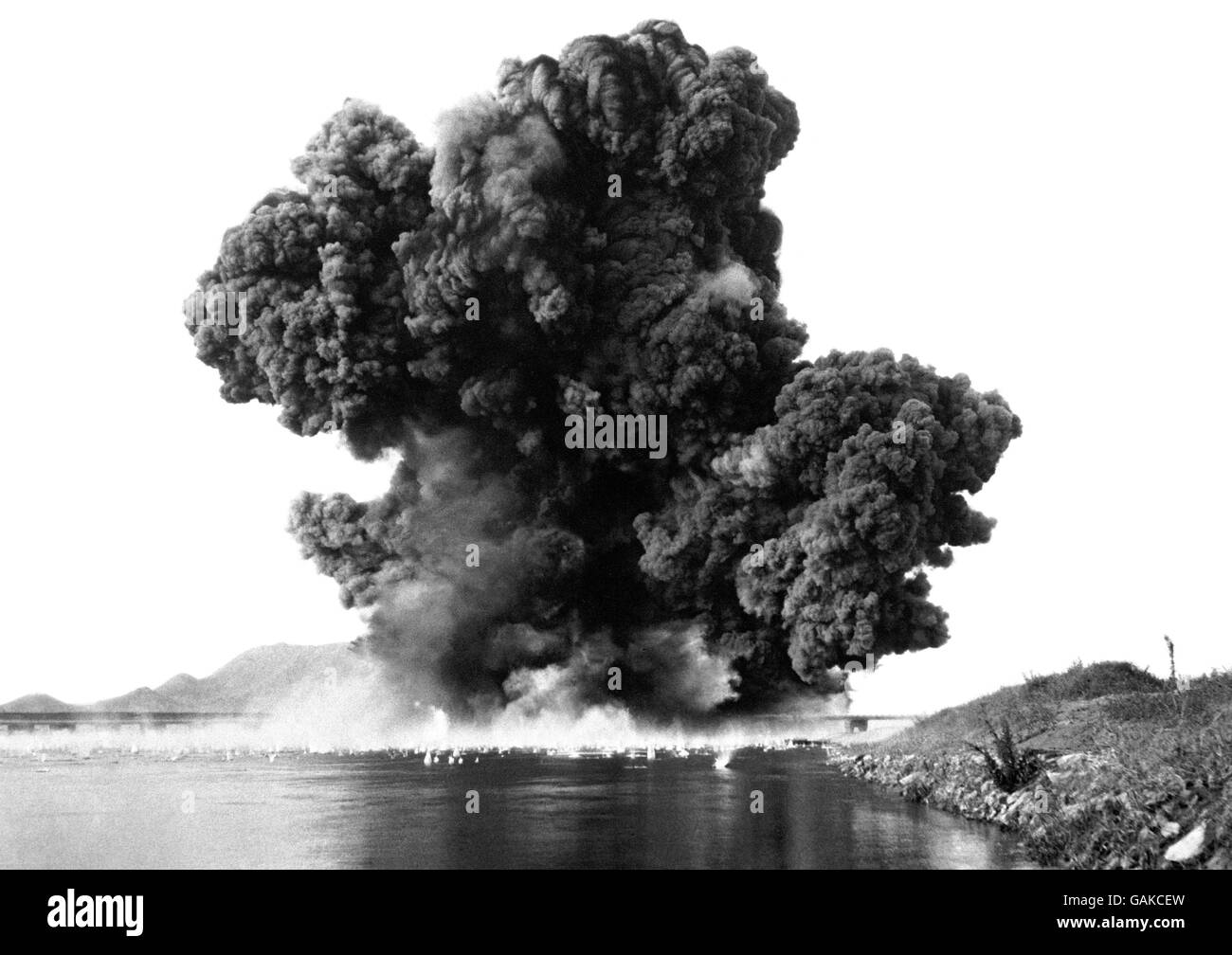 Thick smoke rolls skyward like a dark fleecy cloud, plumes of water rise from the debris - spattered Kum river - and another bridge is closed to the North Korean invaders. The bridge was blown up as the Americans, vastly outnumbered, fought grimly to halt the North Korean steamroller moving south. Stock Photo