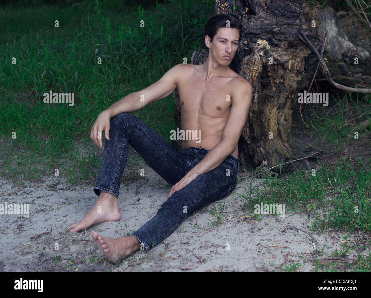 Young man skinny model, fit, sitting on sand grass wood. outdoors leaning on stump sitting sand Stock Photo