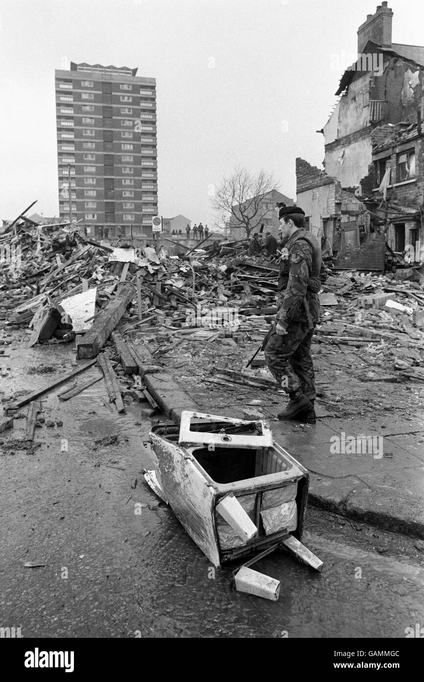 A mass of rubble and, in the foreground, a wrecked gas cooker, as a soldier stands guard over