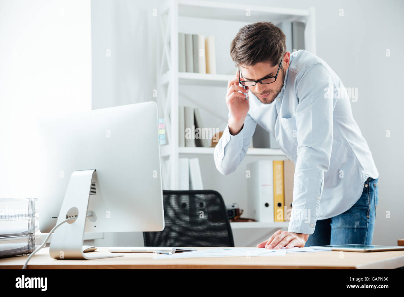 Young handsoome business man standing at desk working on documents with mobile phoone in office Stock Photo