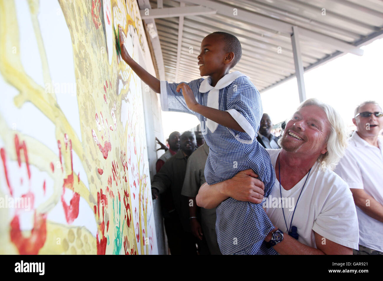 Sir Richard Branson picks up a young school girl so she can hand paint a wall of a new Masi Mara School building in Kenya. Stock Photo