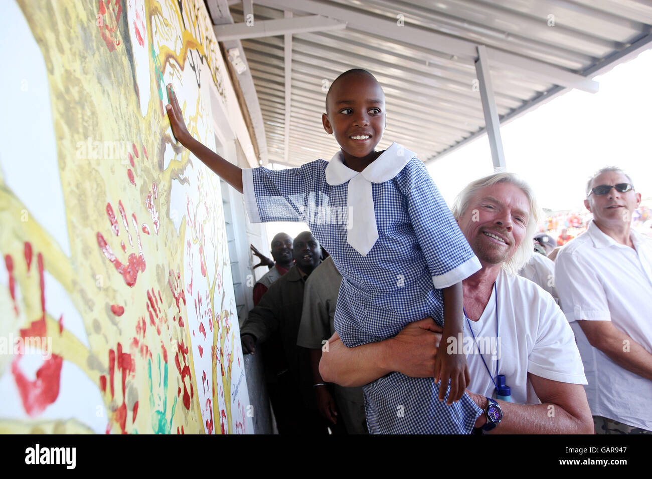 Sir Richard Branson picks up a young school girl so she can hand paint a wall of a new Masi Mara School building in Kenya. Stock Photo