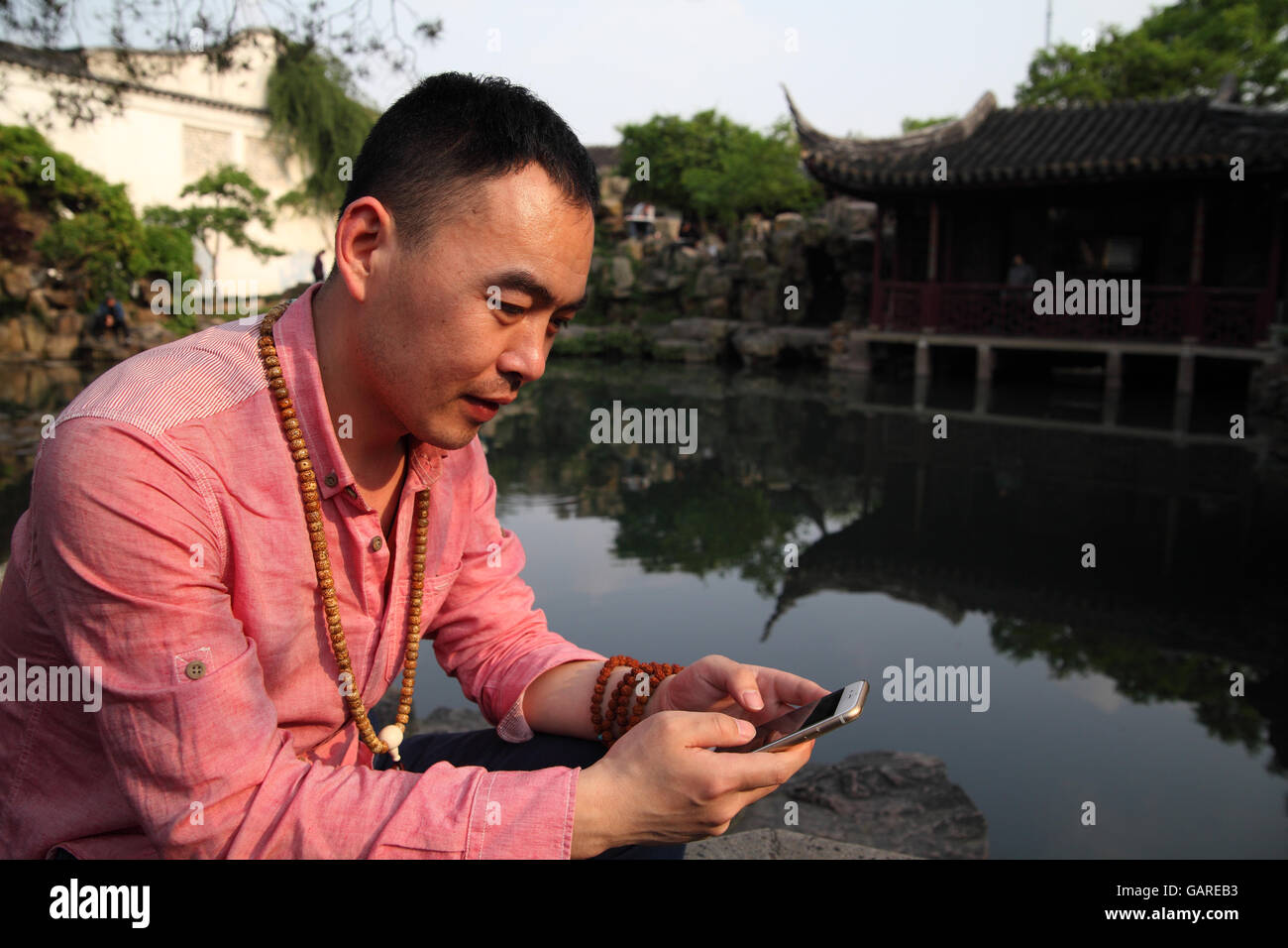 A young Chinese man smiles while playing with his smartphone while sitting by a pond in the Master of Nets Garden. Suzhou, China Stock Photo