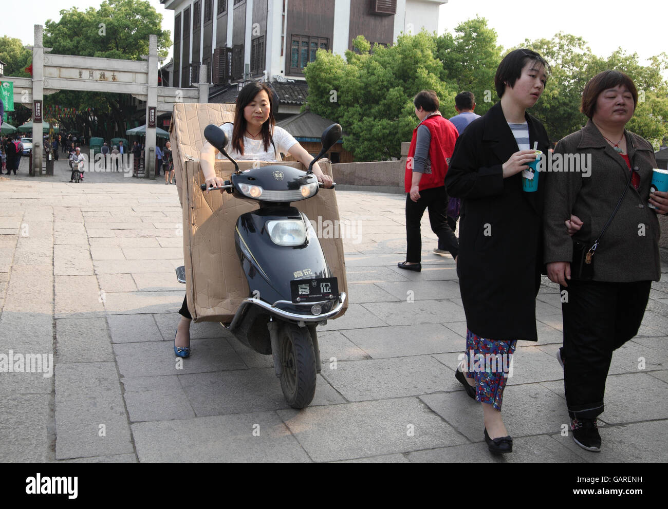 A Chinese woman rides a scooter transporting huge packages she carries in front of her and in the back, other people walk around. Tongli, China. Stock Photo