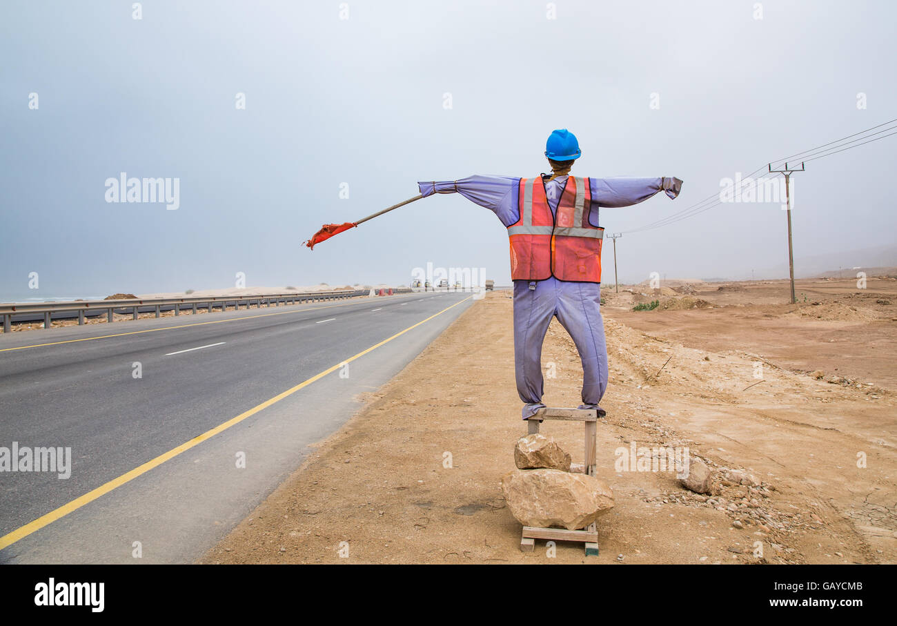 mannequin dressed in safety clothing with a red warning flag standing on the side of the road in Salalah Oman Stock Photo