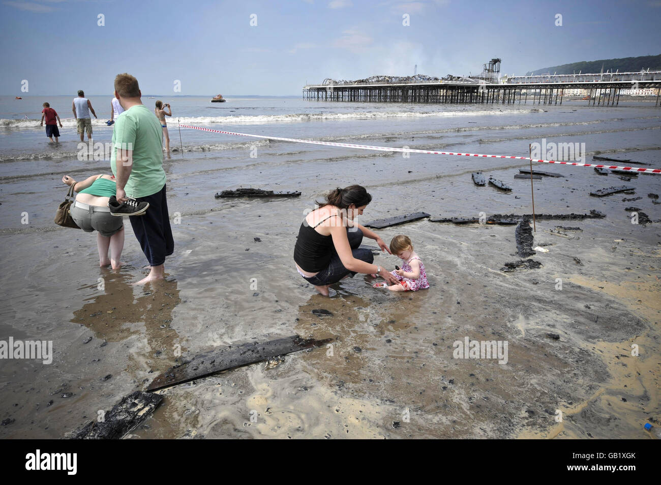 Holiday makers look on as burned debris is washed up on the beach next to the Grand Pier at Weston-super-Mare after a major fire broke out. Stock Photo