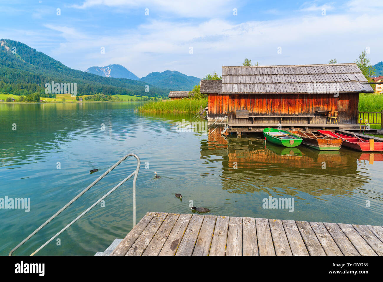 Wooden jetty and fishing boats with wooden houses on shore of Weissensee lake in summer landscape of Carinthia land, Austria Stock Photo