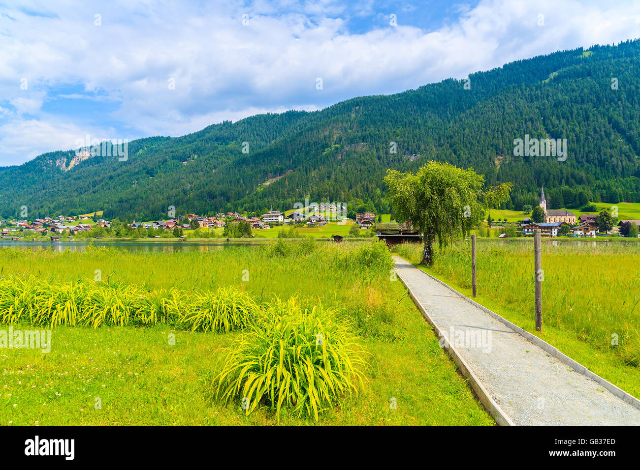 Road along green meadow with alpine village in background in summer landscape of Alps Mountains, Weissensee lake, Austria Stock Photo