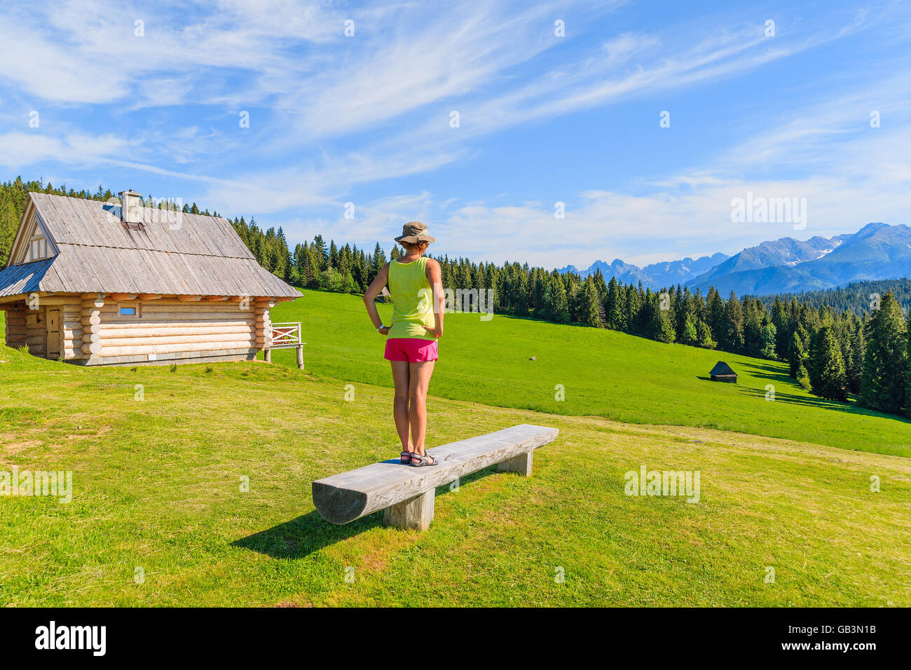 Young woman tourist standing on bench on green meadow and looking at Tatra Mountains, Poland Stock Photo