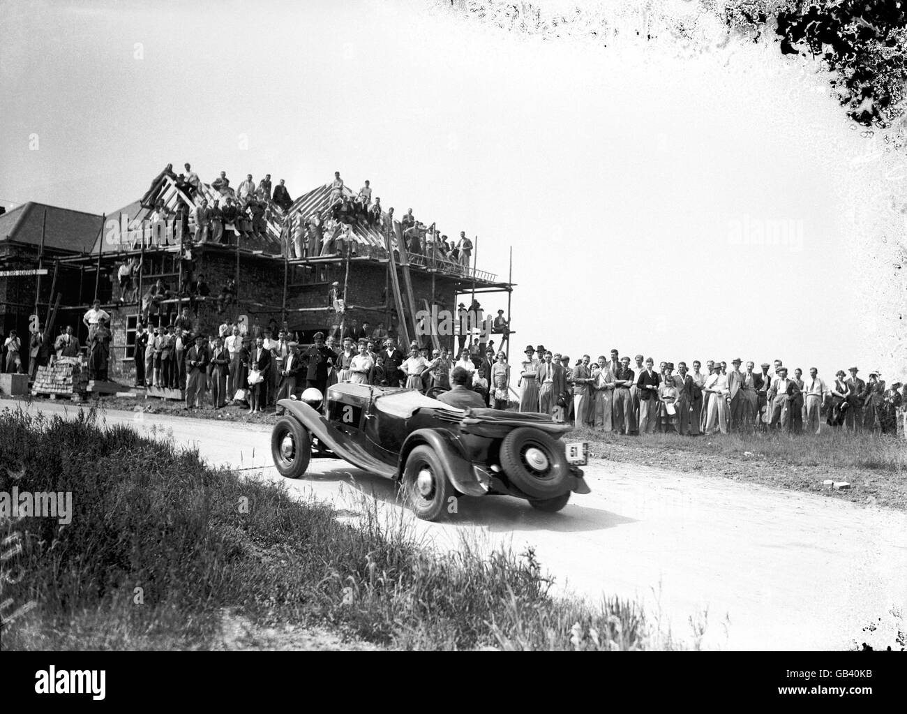 A large crowd watches from the scaffolding and roof-beams of an unfinished house as RRK Marker drives past in his Railton Stock Photo