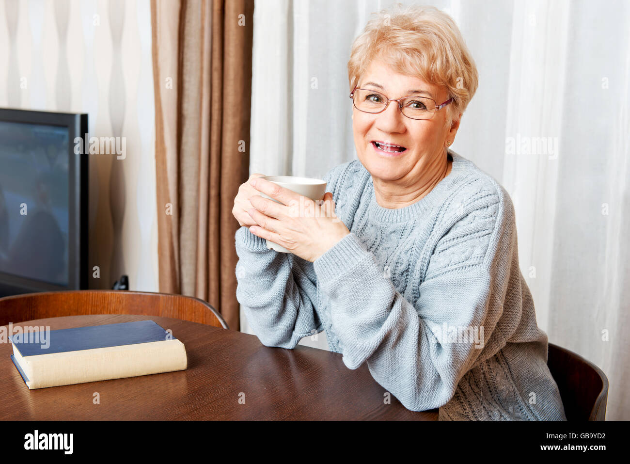 Senior woman sitting at the desk with book and drinking tea or coffee Stock Photo