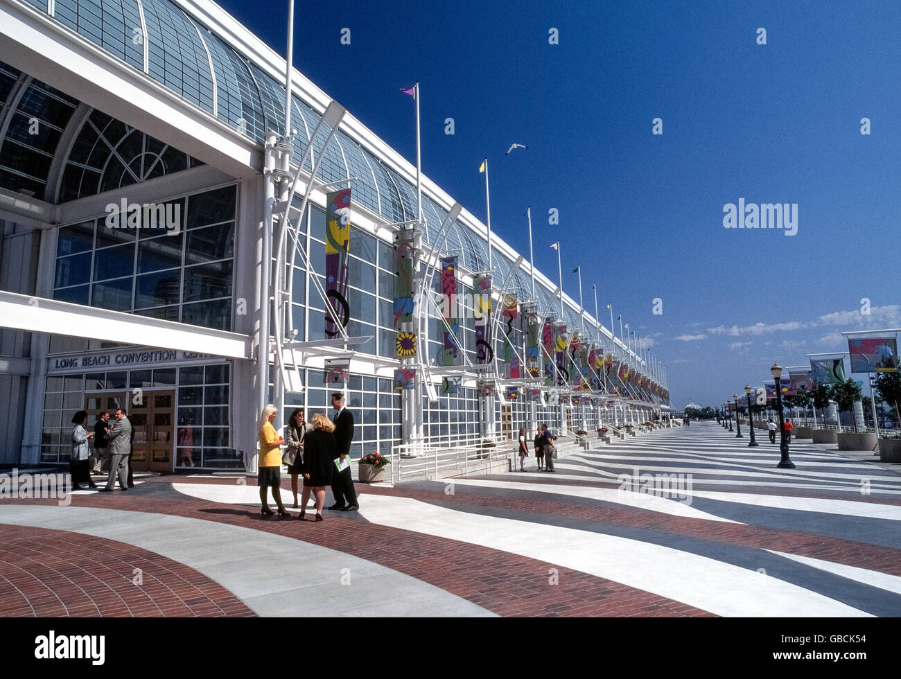 A wide outdoor promenade stretches in front of the glass facade that lets plenty of sunshine into the Long Beach Convention Center located along the downtown waterfront of Long Beach, California, USA. The impressive building holds exhibition halls and meeting rooms that are part of a convention and entertainment complex that also includes two theaters and an arena for concerts and sporting events. Stock Photo