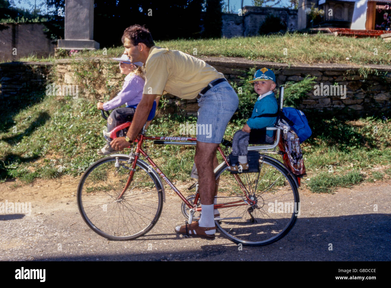 A father is riding a bicycle with two children, the children are sitting in a child seat Stock Photo