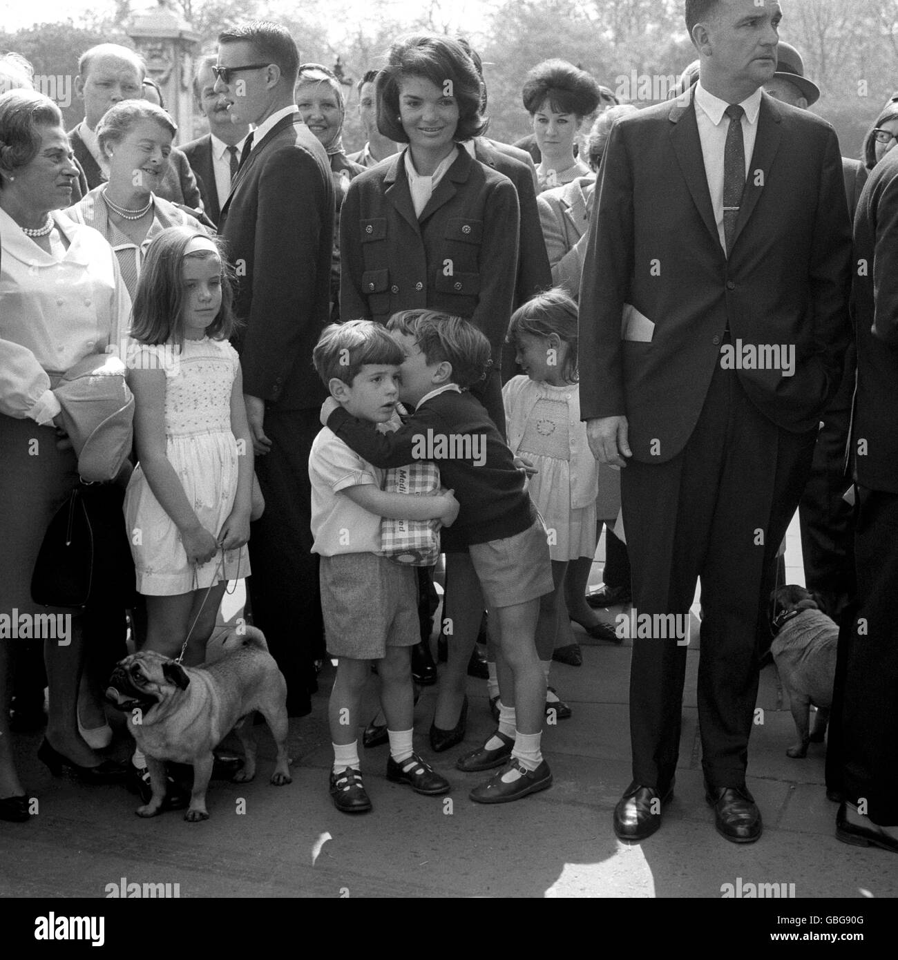 John Kennedy, son of the late American President John F Kennedy, clutches a loaf to his chest and listens as his cousin Anthony Radziwill takes him into his confidence outside the railings of Buckingham Palace forecourt. They were watching the Changing of the Guards before feeding the ducks in St James's Park. Looking on are Jacqueline Kennedy and her daughter Caroline. Stock Photo