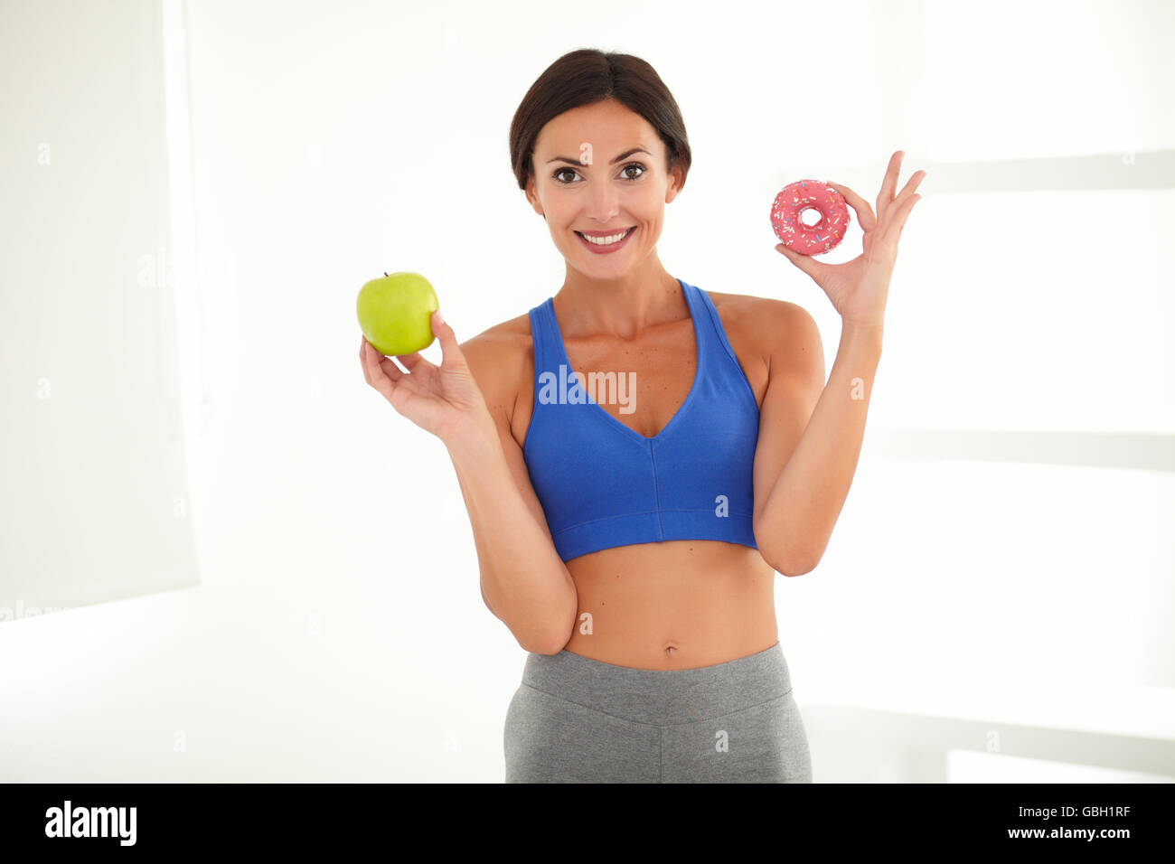 Smiling sporty lady on a diet deciding between apple and sugary donut while looking at you Stock Photo