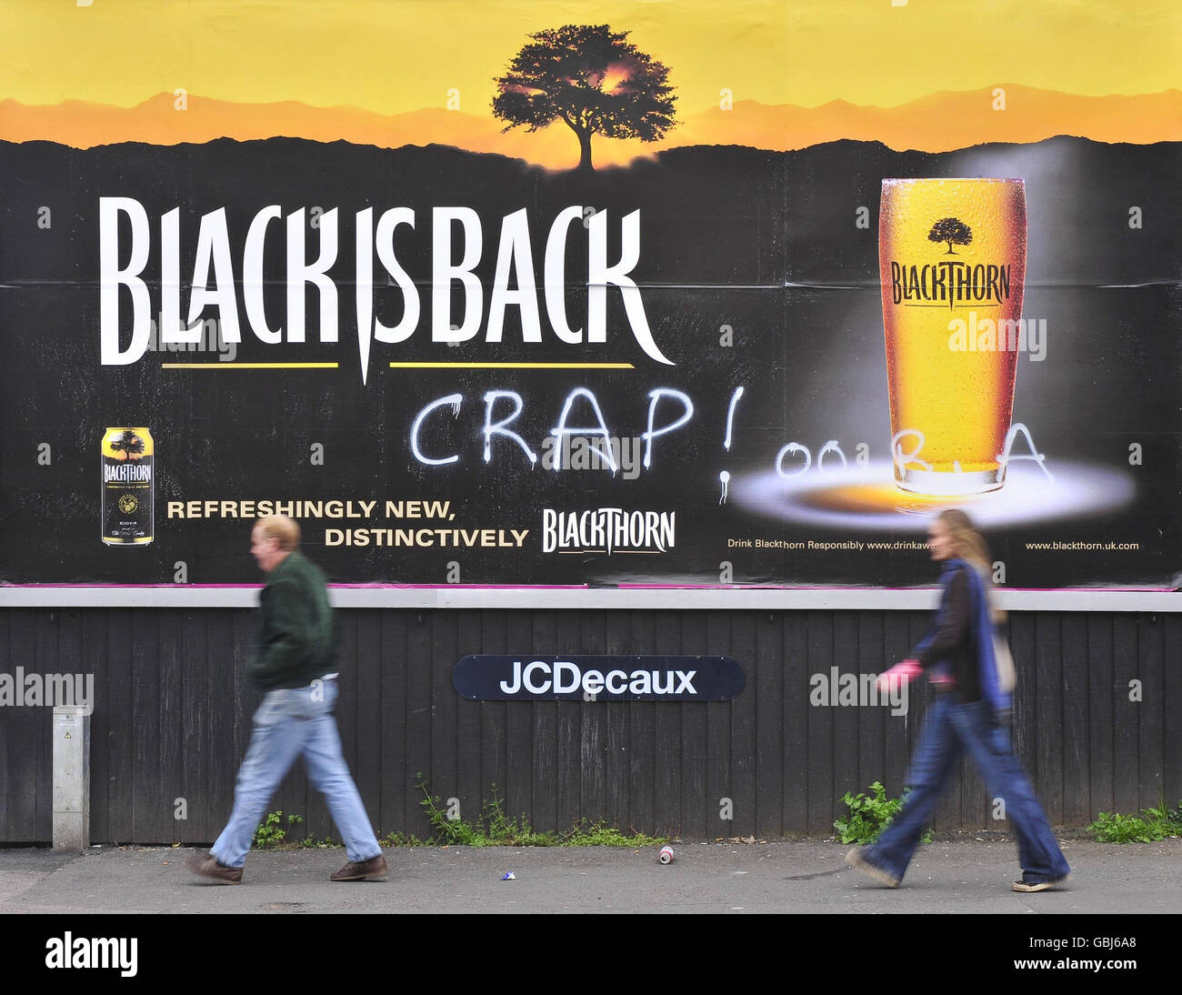 Two people walk past a defaced poster for Blackthorn cider on West Street, Bedminster, Bristol, where at least two posters have been defaced in the area. Stock Photo