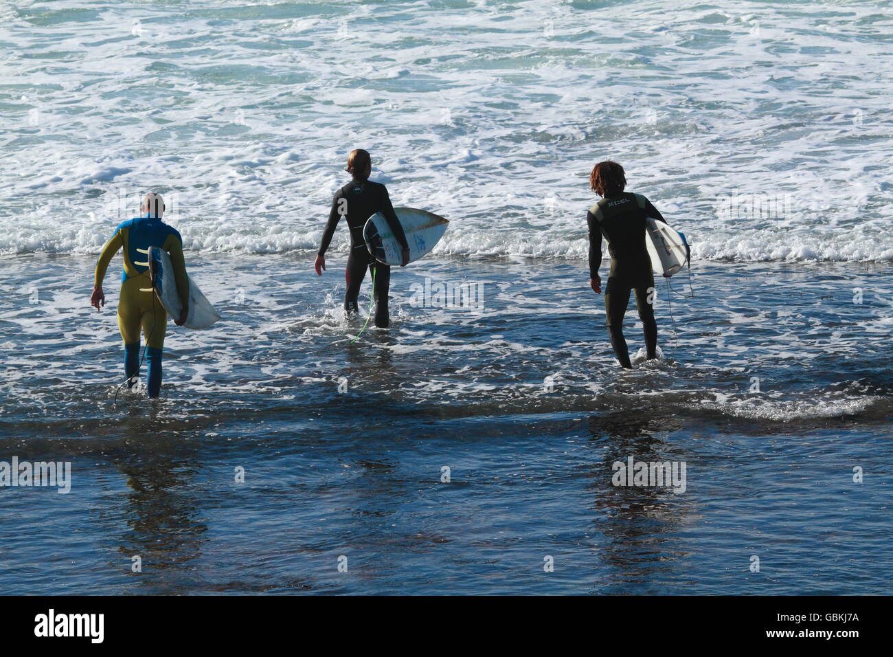 Three male surfers wading into the water in their wetsuits to go