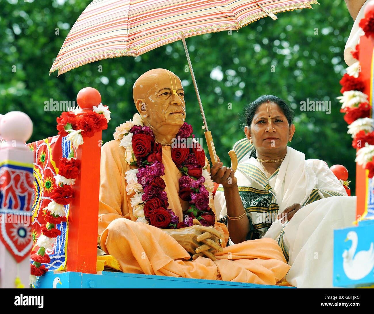 A Hare Krishna devotee keeps the sun off a wooden statue on a giant decorative wheeled shrine, one of three that was hand pulled from Hyde Park to Trafalgar Square to celebrate the 'Ratha-yatra' Festival. Stock Photo