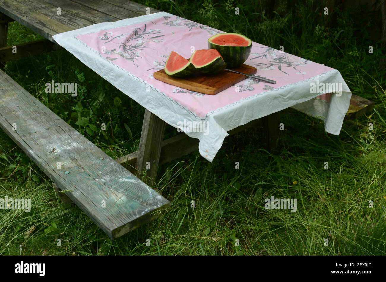 cut watermelon pieces on bamboo cutting board, knife, on vintage tablecloth on wood picnic table in grass Stock Photo