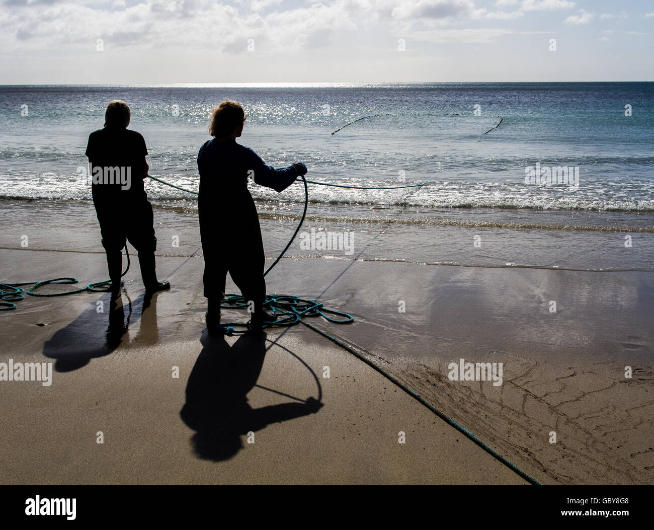 The silhouettes of two Cornish fishermen standing on a beach at sunrise and hauling their nets in as they fish for sand eels. Stock Photo