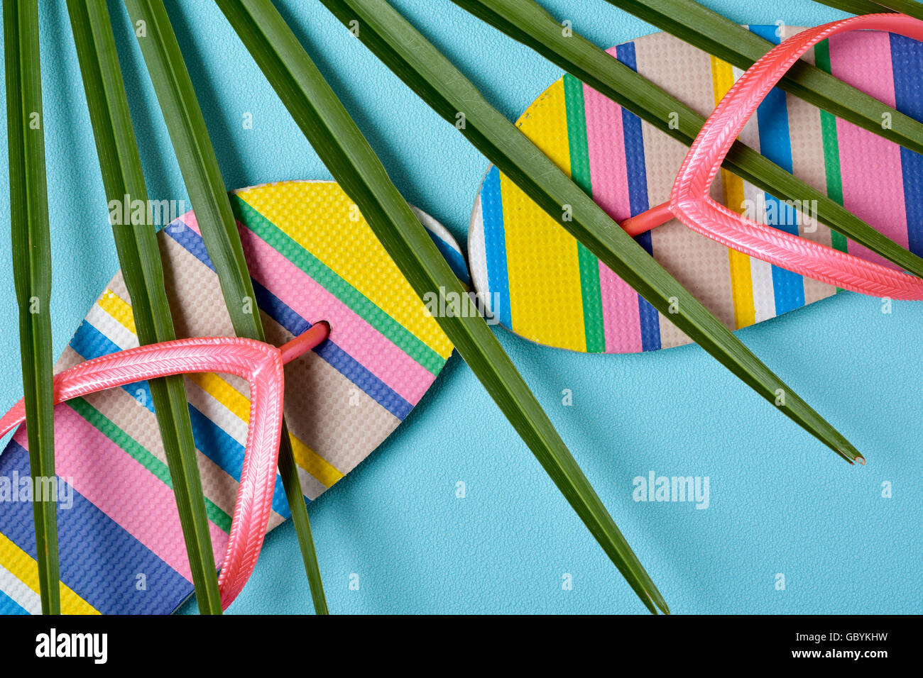 closeup of a pair of colorful striped-patterned flip-flops with pink straps and a palm leaf on a blue background Stock Photo