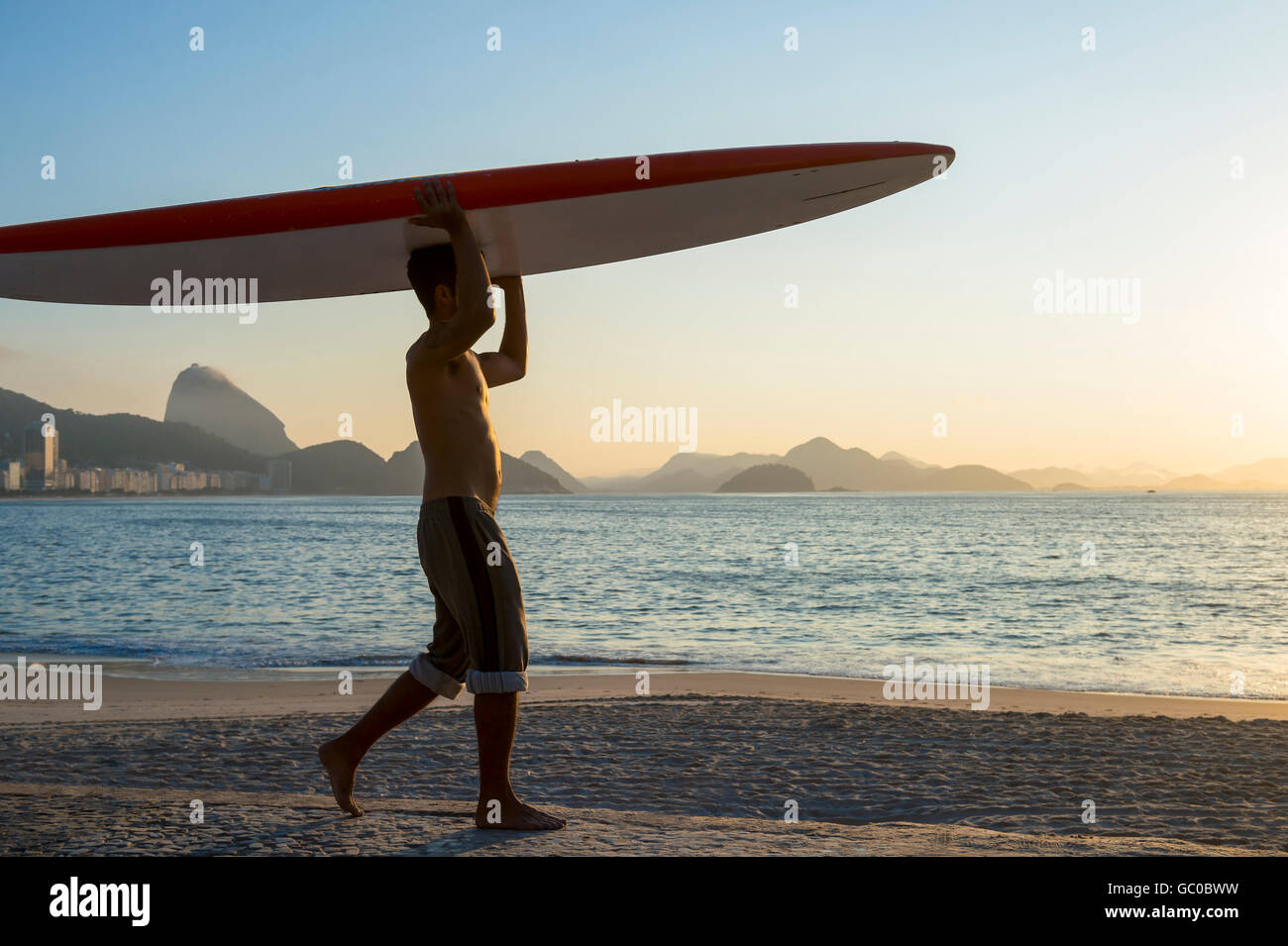 RIO DE JANEIRO - APRIL 5, 2016: A young Brazilian man on Copacabana Beach balances a surfboard on his head at sunrise. Stock Photo