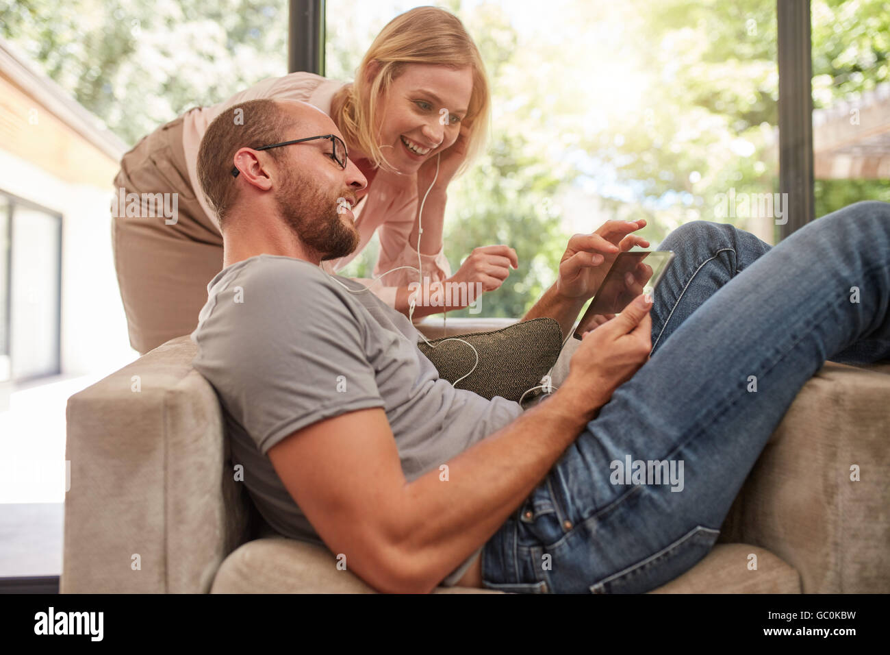 Indoor shot of a  couple listening to music on a digital tablet and sharing earphones. Man lying on sofa with his wife standing Stock Photo