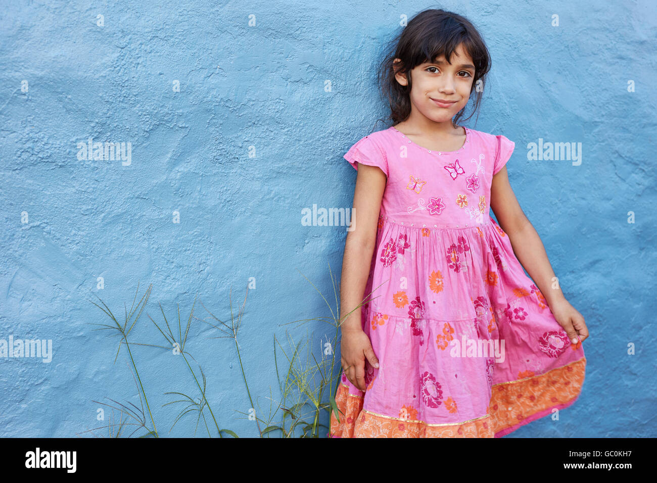 Shot of cute little girl in beautiful pink dress looking at camera while standing against blue wall. Stock Photo