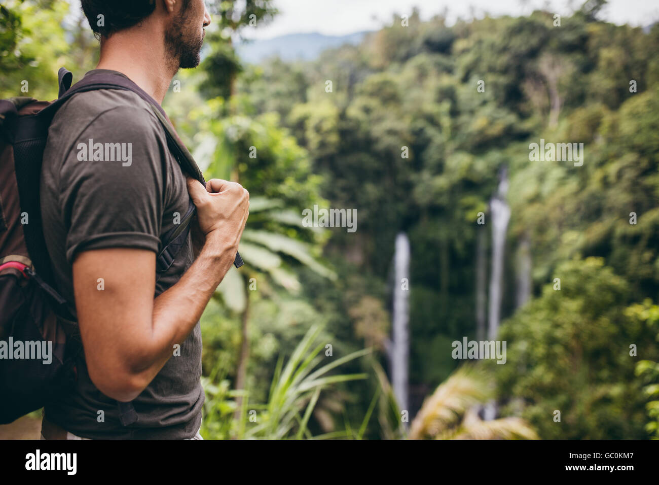 Close up shot of young man with backpack standing in nature and looking at waterfall. Male hiker near waterfall in forest. Stock Photo