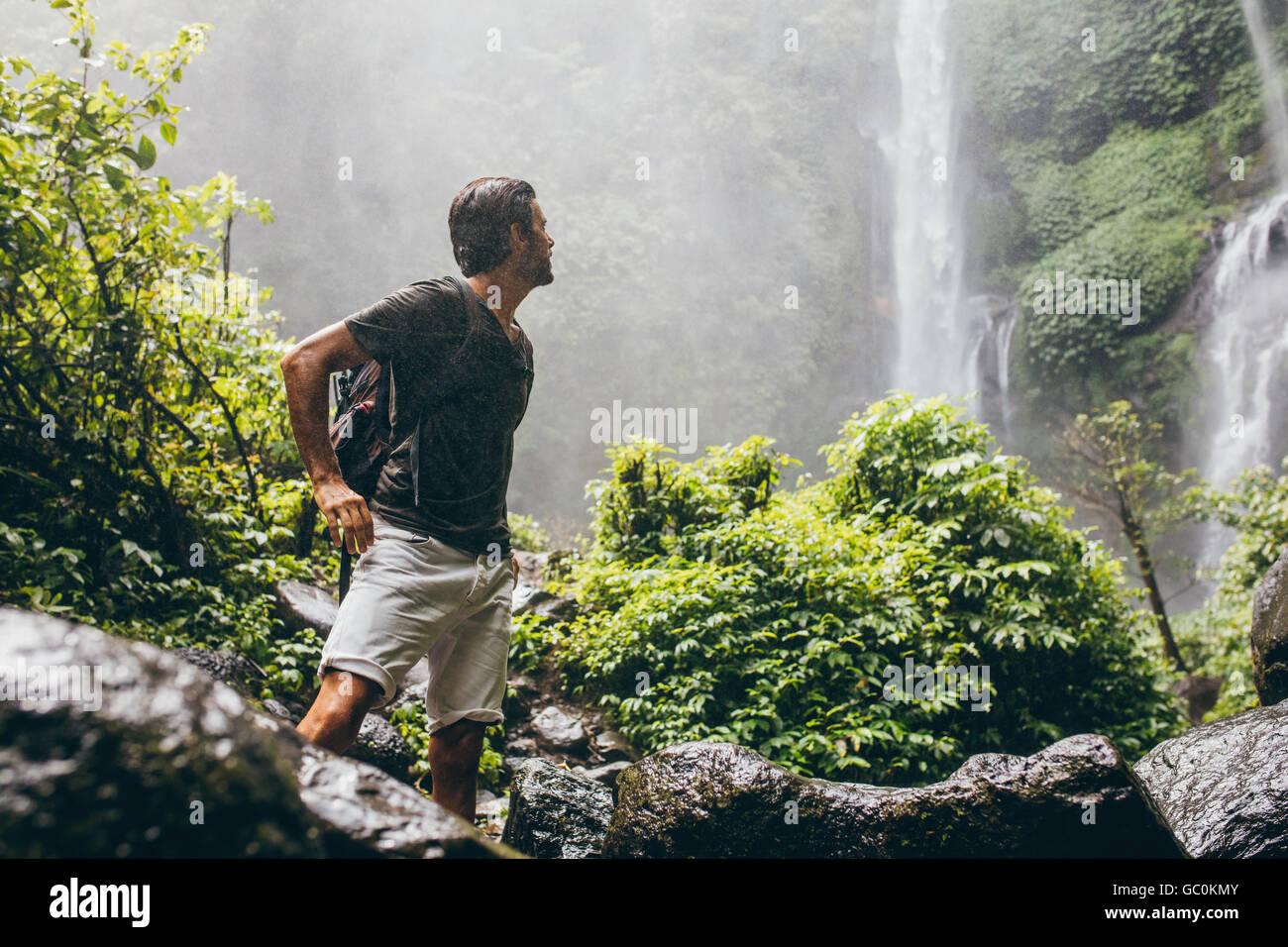 Young man with backpack standing near a waterfall in forest. Male hiker in the nature during rain. Stock Photo