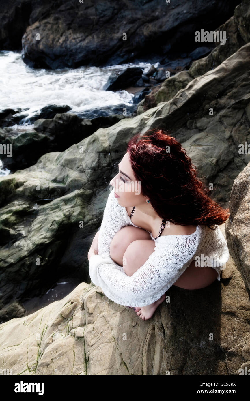 Hispanic Woman Holding Knees To Chest Sitting On Rocks At Sea Shore Looking Out Over The Water In Solitude Stock Photo