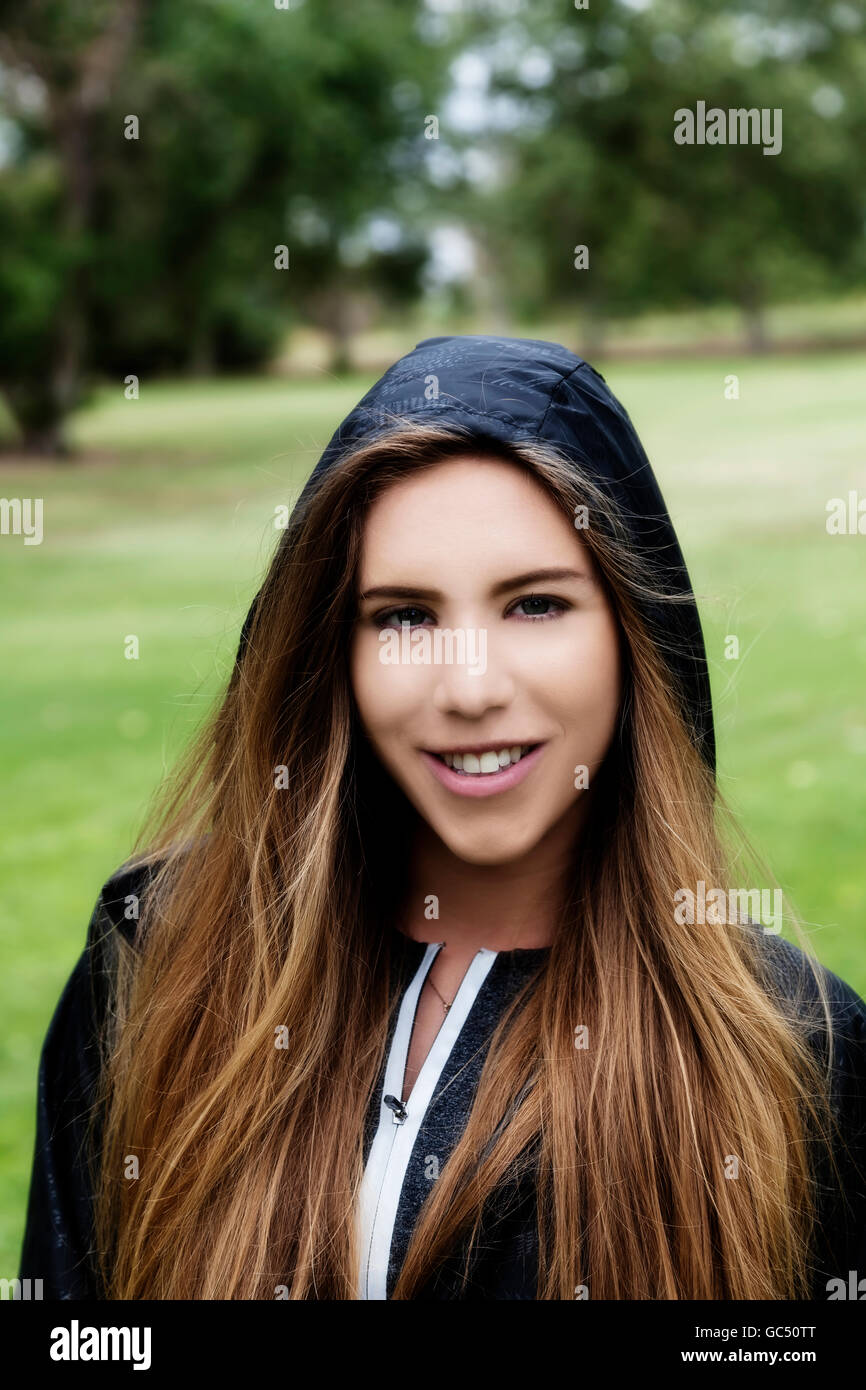 Attractive Smiling Caucasian Teen Woman Outdoor Portrait At Park With Green Grass And Trees Wearing Black Hooded Jacket Stock Photo