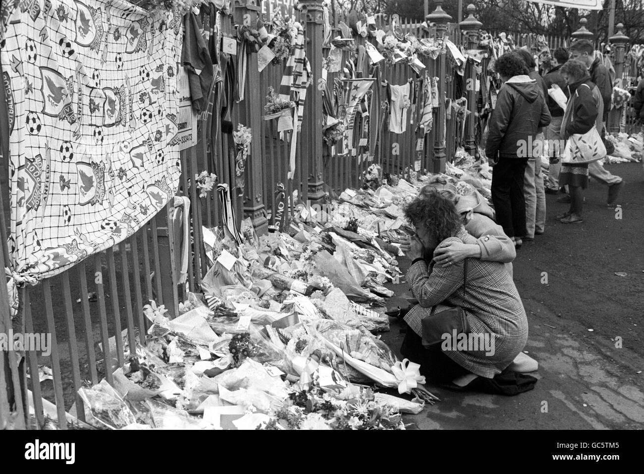 A WOMAN IS COMFORTED AS SHE KNEELS BY FLORAL TRIBUTES AT THE GATE OF THE HILLSBOROUGH GROUND, SHEFFIELD, THE MORNING AFTER NINETY SIX LIVERPOOL FANS DIED FROM INJURIES SUFFERED IN THE FATAL CRUSH AT THE GROUND. Stock Photo