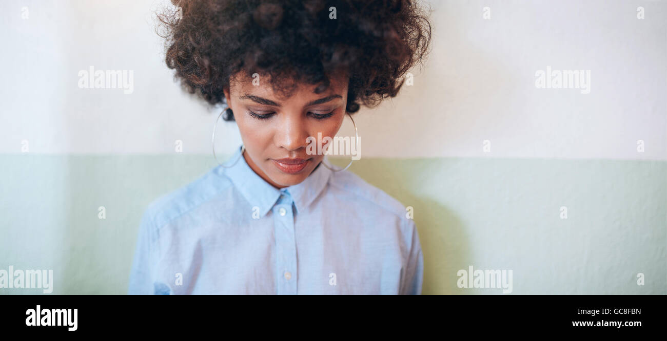 Horizontal shot of young woman with curly hair looking down while standing against a wall. African woman looking pensive. Stock Photo