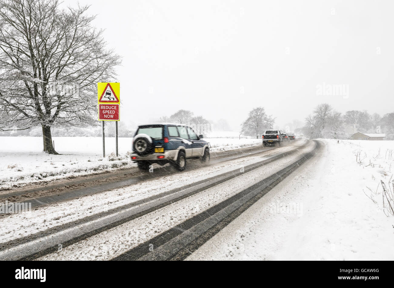 Cars drive past a slippery surface warning road sign while it is snowing with snow on the ground. Stock Photo