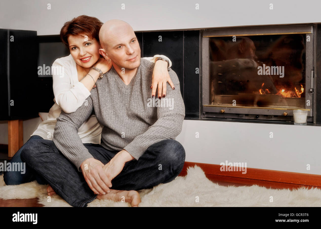 Romantic couple of brunette woman and bald headed man sitting on the fur carpet near the fireplace. Fire in the fireplace is burning. Stock Photo