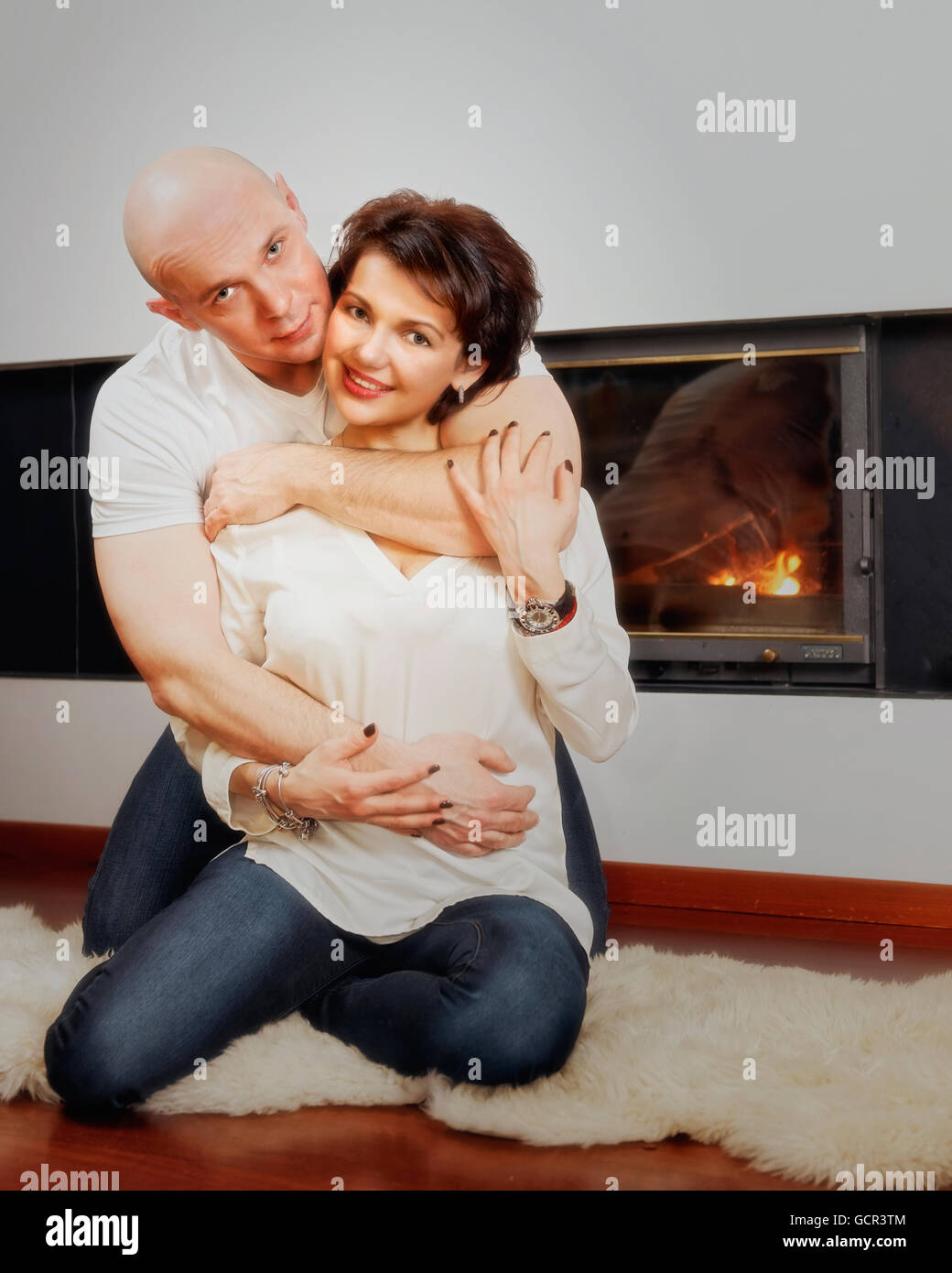 Romantic couple of brunette woman and bald headed man sitting on the fur carpet near fireplace. Fire in the fireplace is burning. Stock Photo