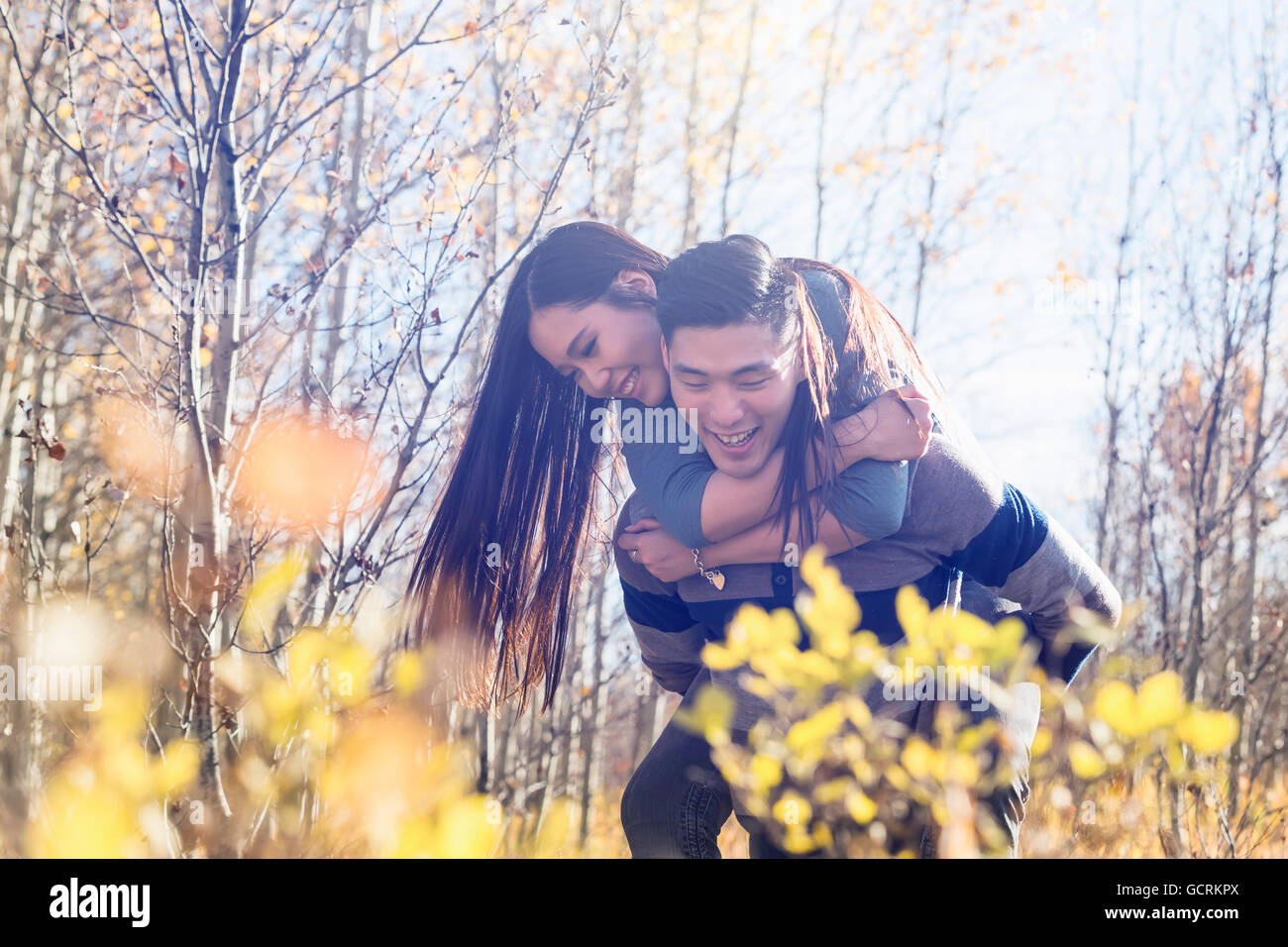 A young Asian woman riding piggy back on her boyfriend while walking in a park in autumn; Edmonton, Alberta, Canada Stock Photo