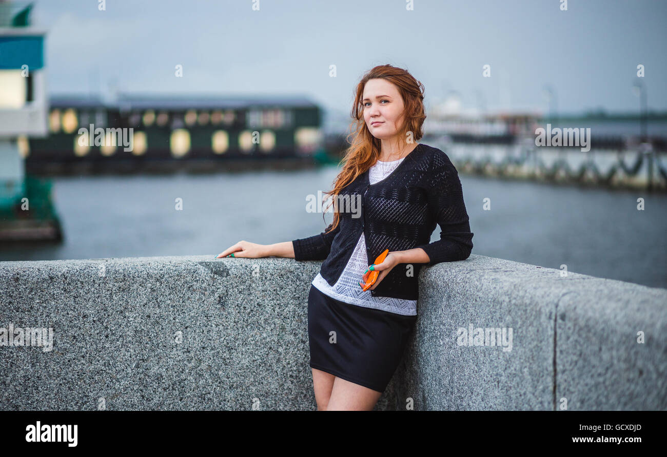Beautiful girl standing on the bank of the river. She holding phone and looking afar. Stock Photo