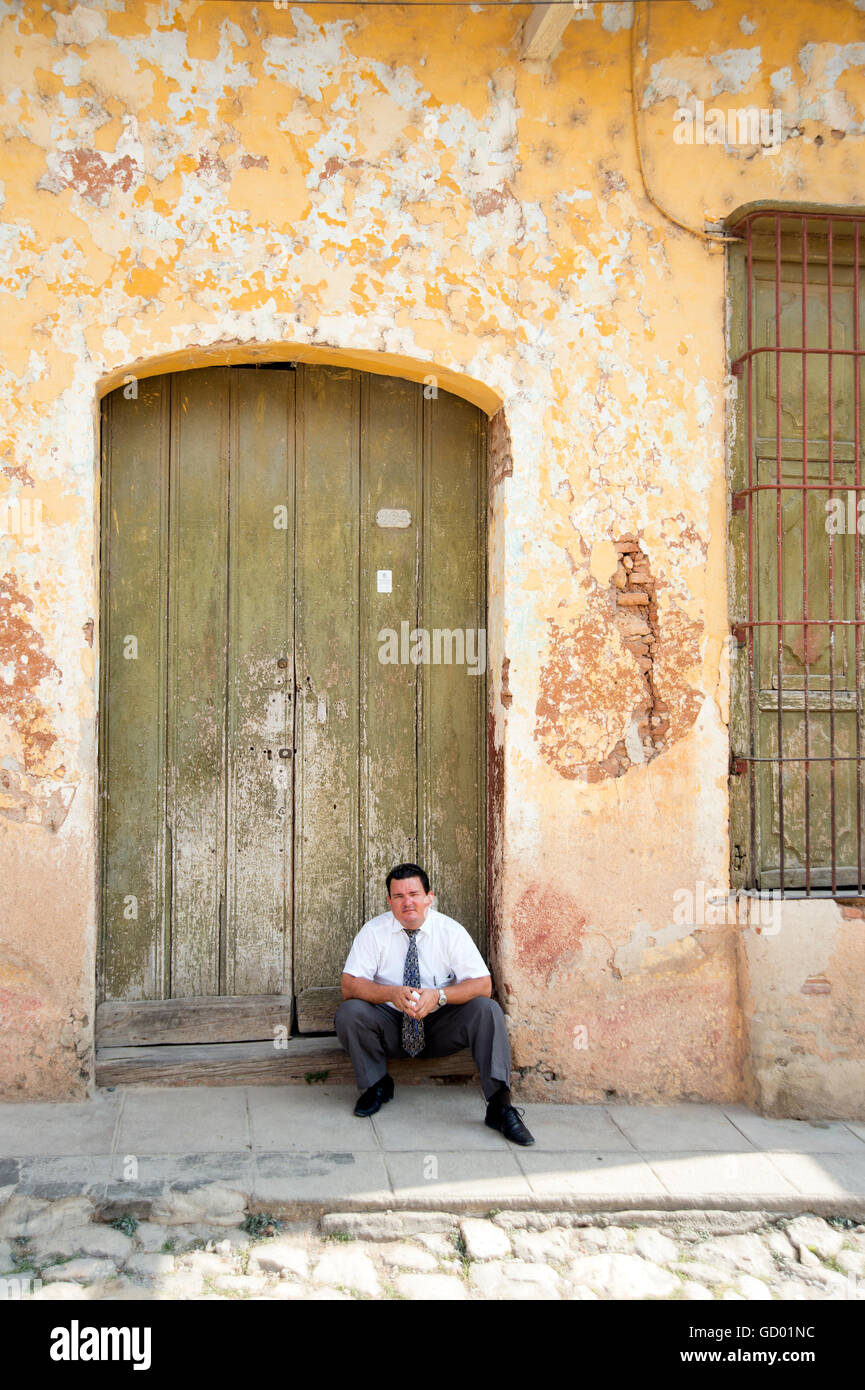 A smart dressed Cuban restaurant doorman sits in the shade of a doorway before the evening trade in Trinidad Cuba Stock Photo