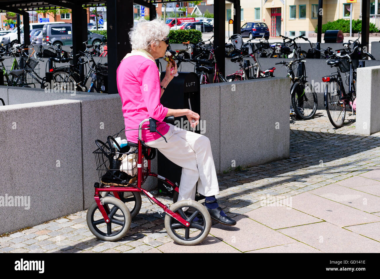 Ronneby, Sweden - July 9, 2016: Senior woman sitting on walker eating ice cream. Parking lot for bikes in the background. Stock Photo