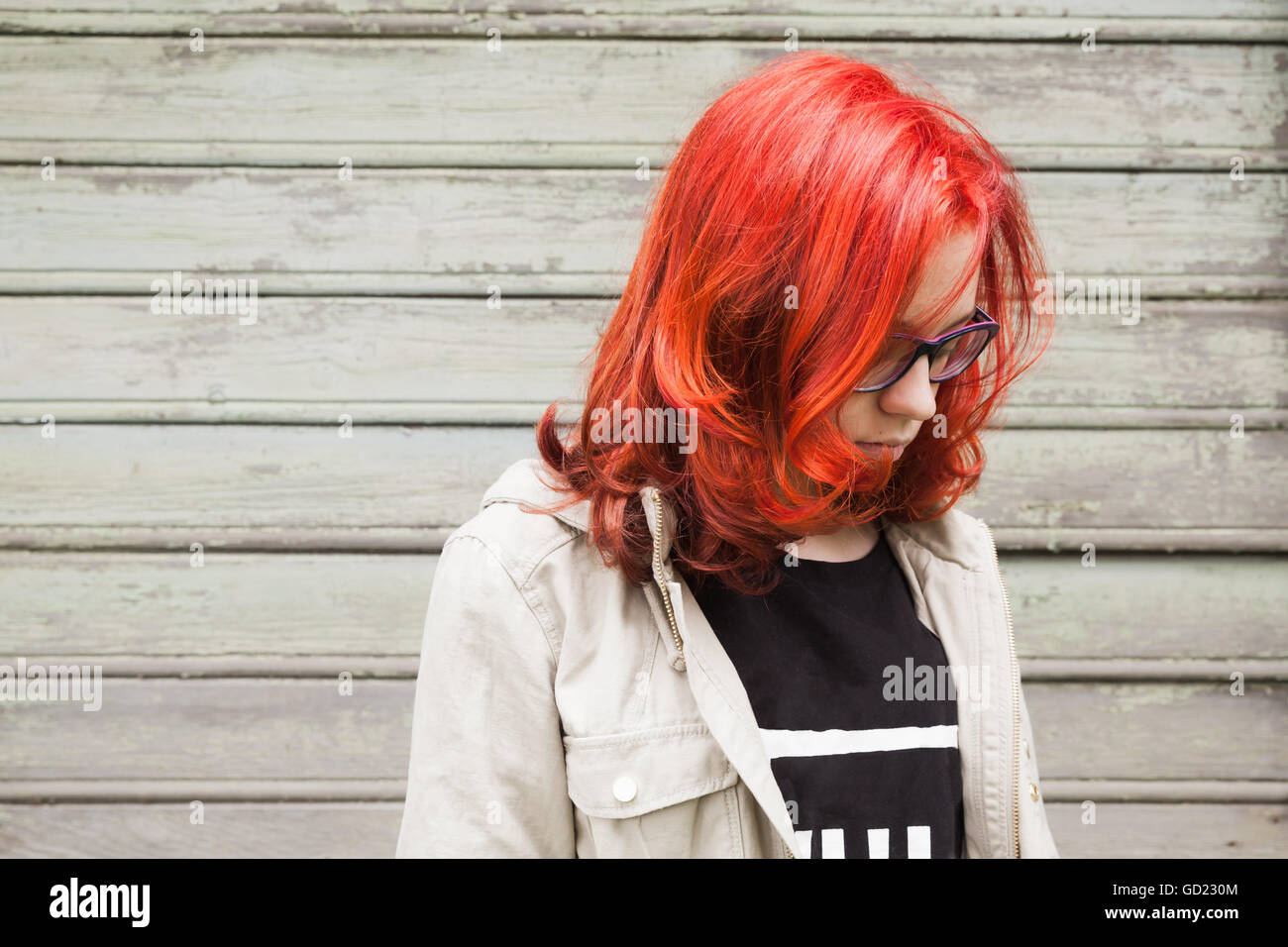 Sad Caucasian teenager girl in glasses with bright red hair, closeup outdoor portrait over green grungy wooden wall Stock Photo