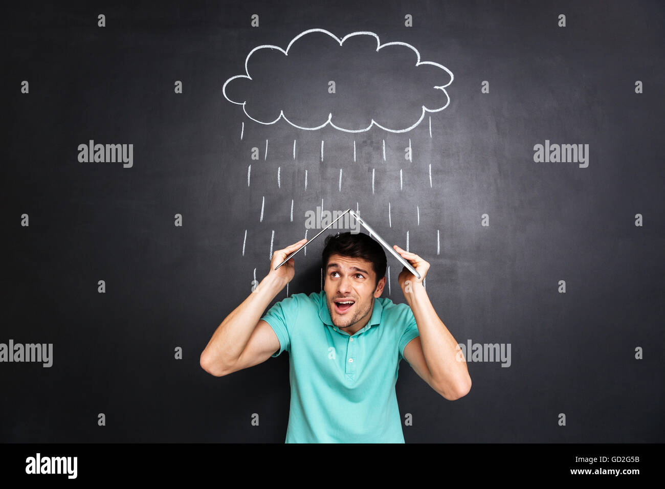 Stressed frowning young man holding laptop above head and covering from drawn rain over blackboard background Stock Photo