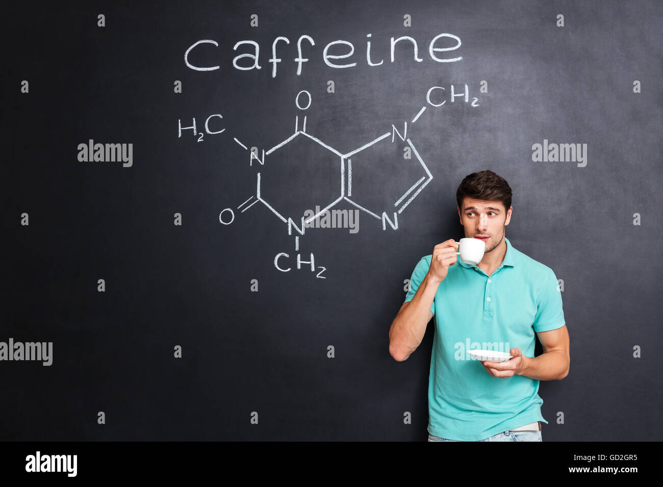 Handsome young man drinking coffee over blackboard background with drawn chemical structure of caffeine molecule Stock Photo